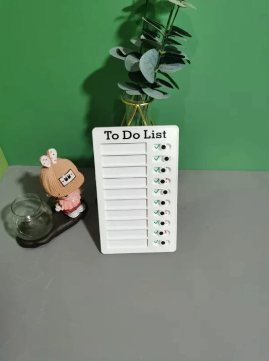 White reusable to-do list board with sliding check marks, styled on a table next to a doll, glass jar, and green plant against a green background.