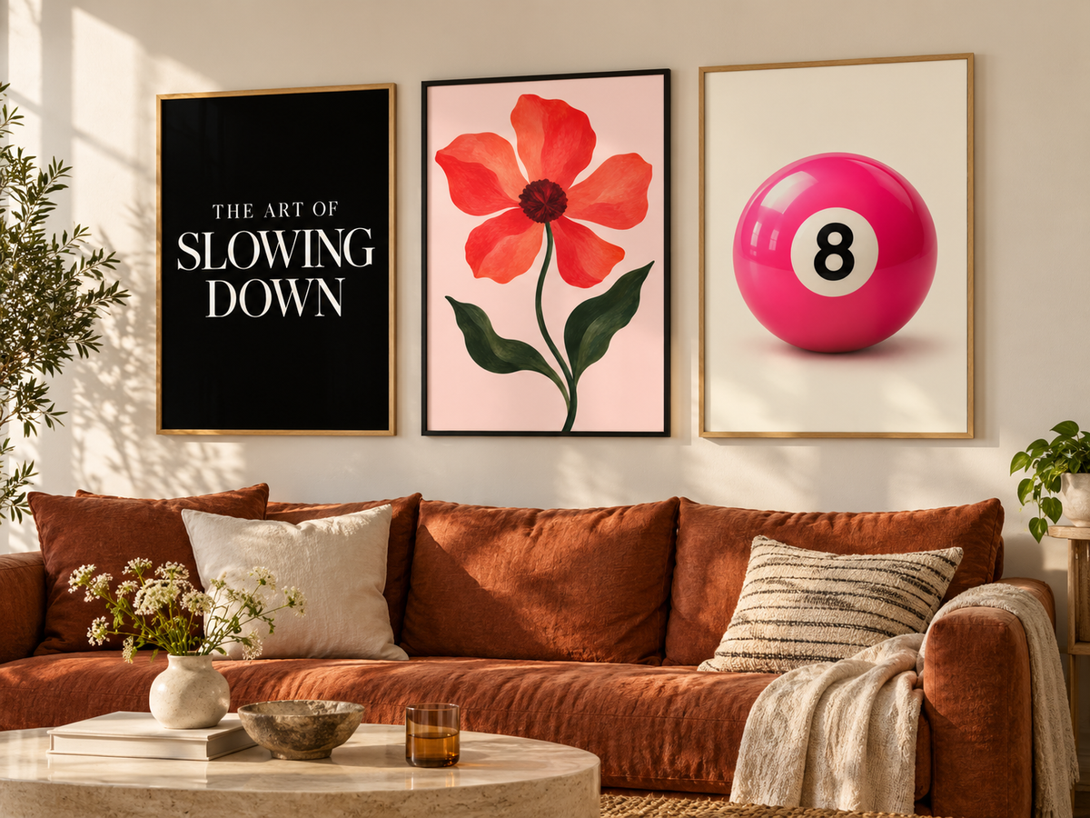 Three framed wall art prints hang above a rust-orange sofa in a styled living room. The left print is black with white text reading 'THE ART OF SLOWING DOWN,' the center is a pink floral artwork with green leaves, and the right shows a bright pink billiard ball with the number 8.
