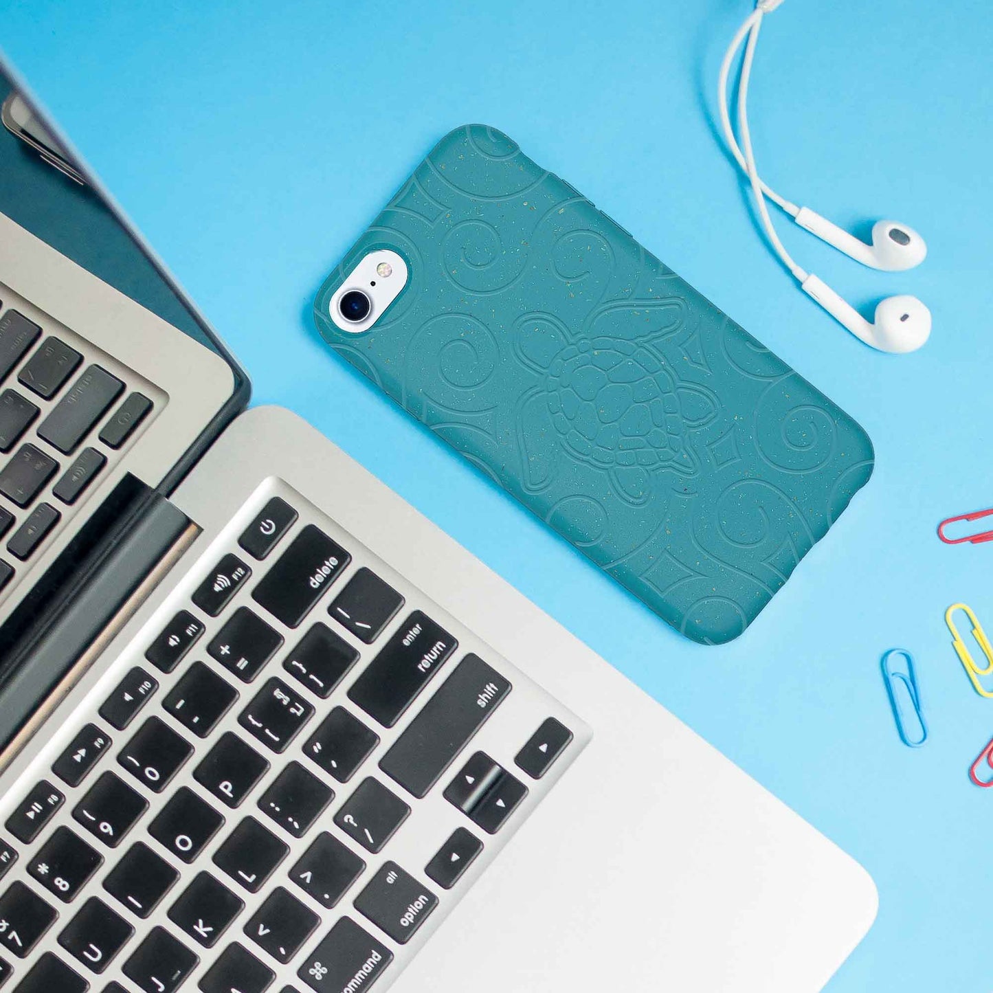 Teal phone case on a laptop with earphones and paper clips on a blue background