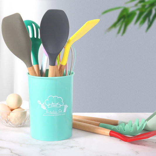 Colorful kitchen utensils in a turquoise holder on a marble surface with a plant in the background.