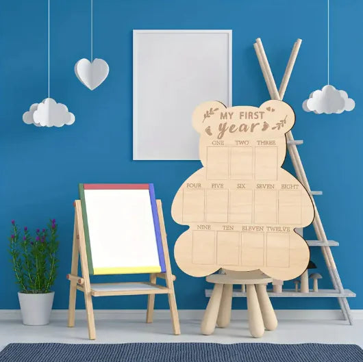 Wooden teddy bear-shaped milestone board with 'My First Year' text in a room with blue walls, a plant, and an easel.