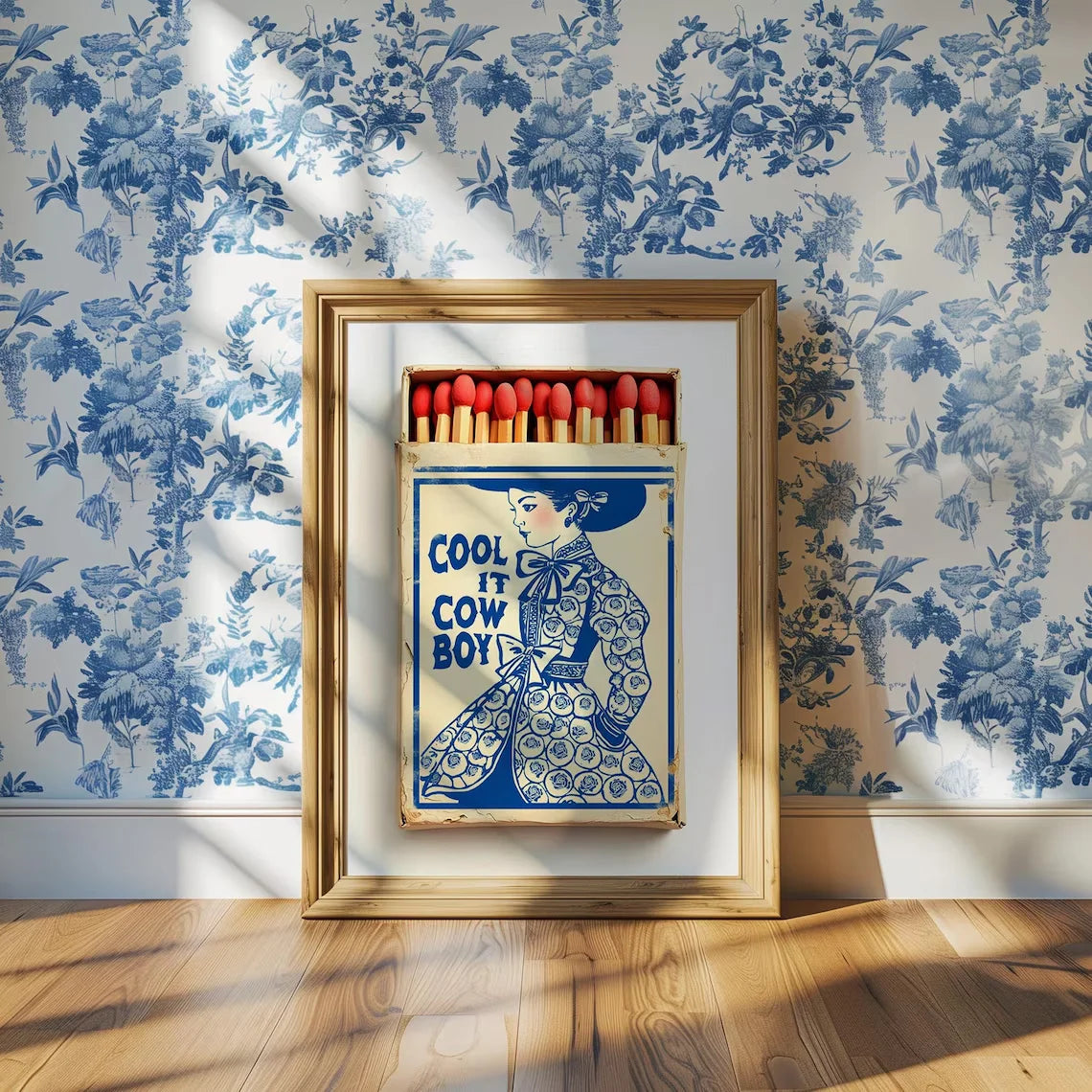 Framed artwork with a matchbox design featuring a woman and text, against a floral wallpapered wall.