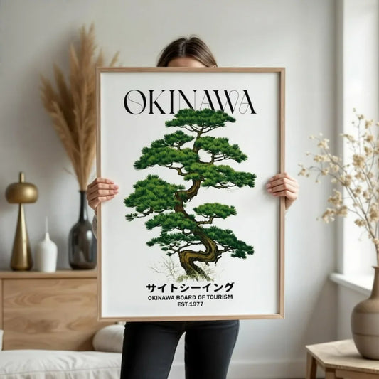 Person holding a framed poster of a green tree with 'Okinawa' text in a room.
