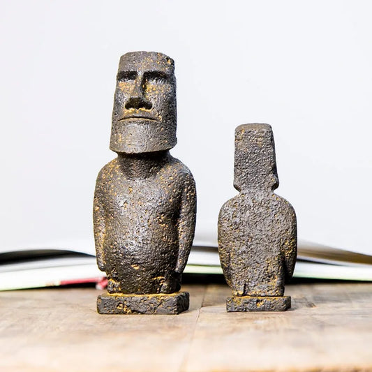Two small Easter Island moai statue figurines, one larger facing forward and one smaller turned away, displayed on a wooden surface with a blurred book in the background.