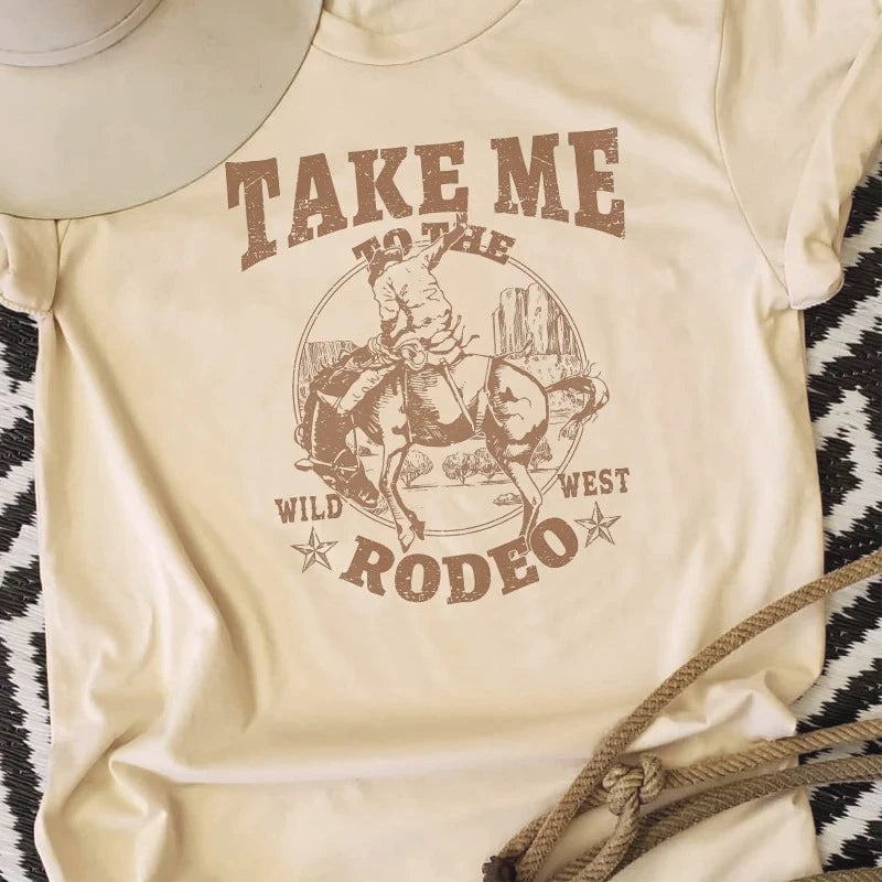 Beige t-shirt with a graphic of a person riding a horse and text 'Take Me to the Wild West Rodeo' on a patterned surface.