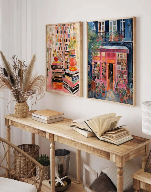 Two framed colorful paintings hanging on a white wall above a wooden console table. The left painting shows stacks of books with warm pink, orange, and cream tones, and the right painting depicts a vibrant storefront scene in pink, blue, and green tones.