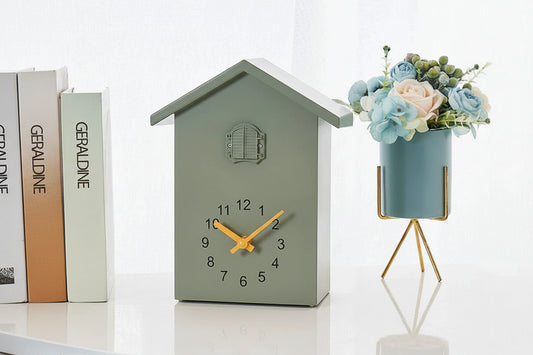 Green house-shaped clock with yellow hands next to a small plant on a white background