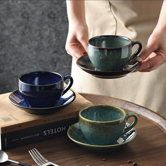 Person holding a green ceramic cup and saucer with two other cups on a wooden table.