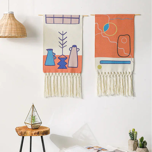 Two colorful wall hangings with abstract designs on a white wall, with a wooden table and plants in the foreground.