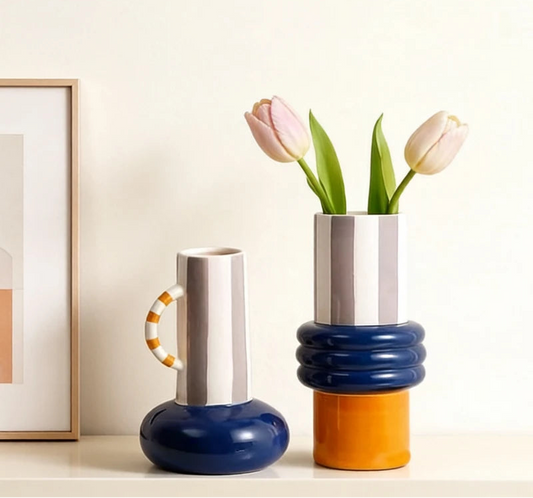 Two modern stacked ceramic vases on a white shelf against a cream wall, with one vase holding two tulips. The vases feature bold color-blocked designs in navy blue, white, orange, and navy, with one vase including a striped orange-and-white handle.