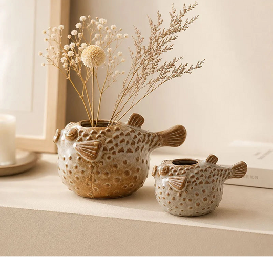Two fish-shaped ceramic vases with a textured, spotted pattern in beige and brown, one filled with dried flowers, displayed on a shelf.