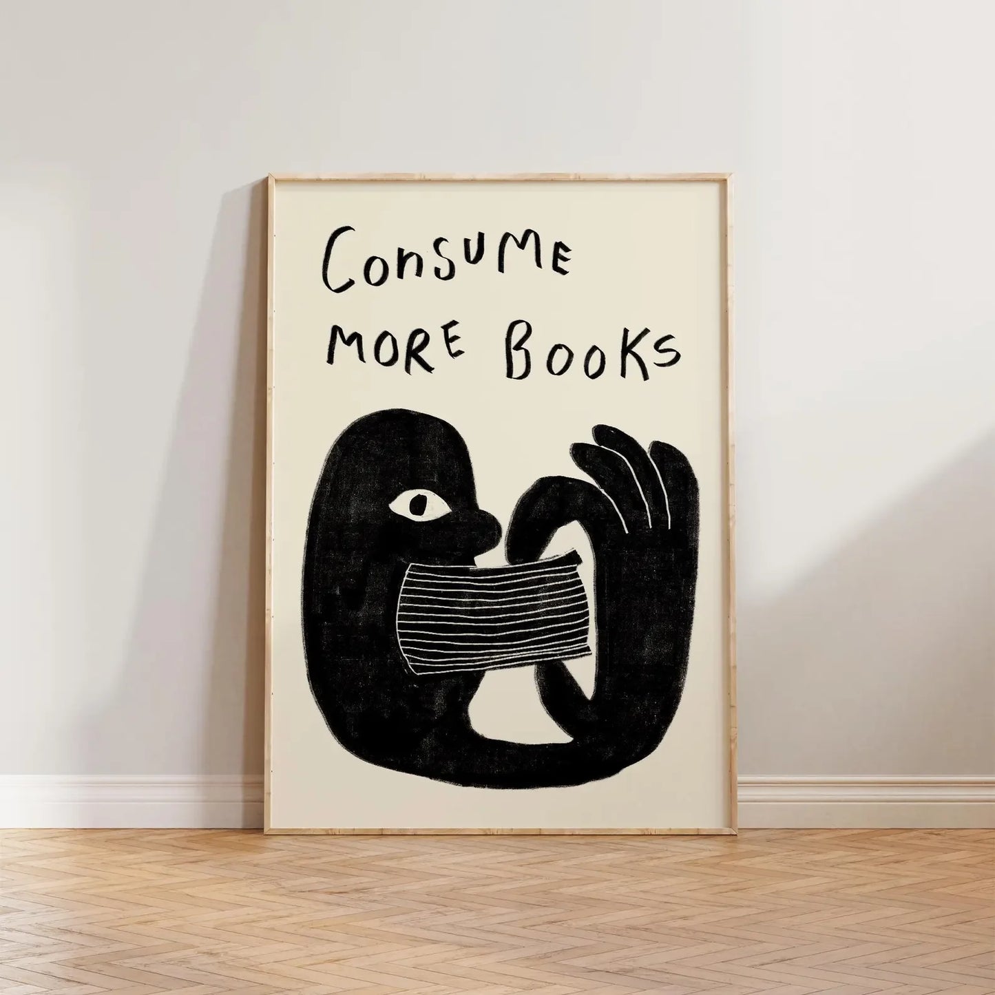 Framed artwork with a black and white illustration of a hand holding a book and text 'CONSUME MORE BOOKS' on a beige background.