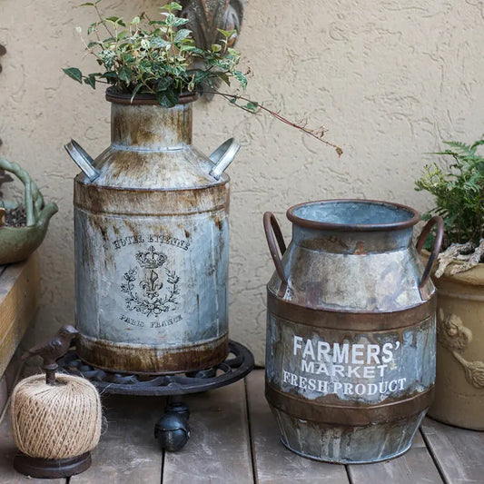 Two rustic metal milk churns with 'Farmers' Market Fresh Product' text on a wooden surface.