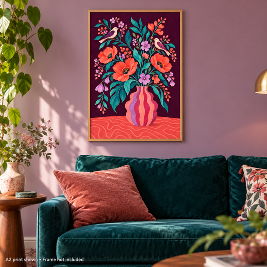A framed art print featuring a colorful floral bouquet in a striped vase with birds, displayed on a mauve wall above a dark teal velvet sofa in a styled living room.