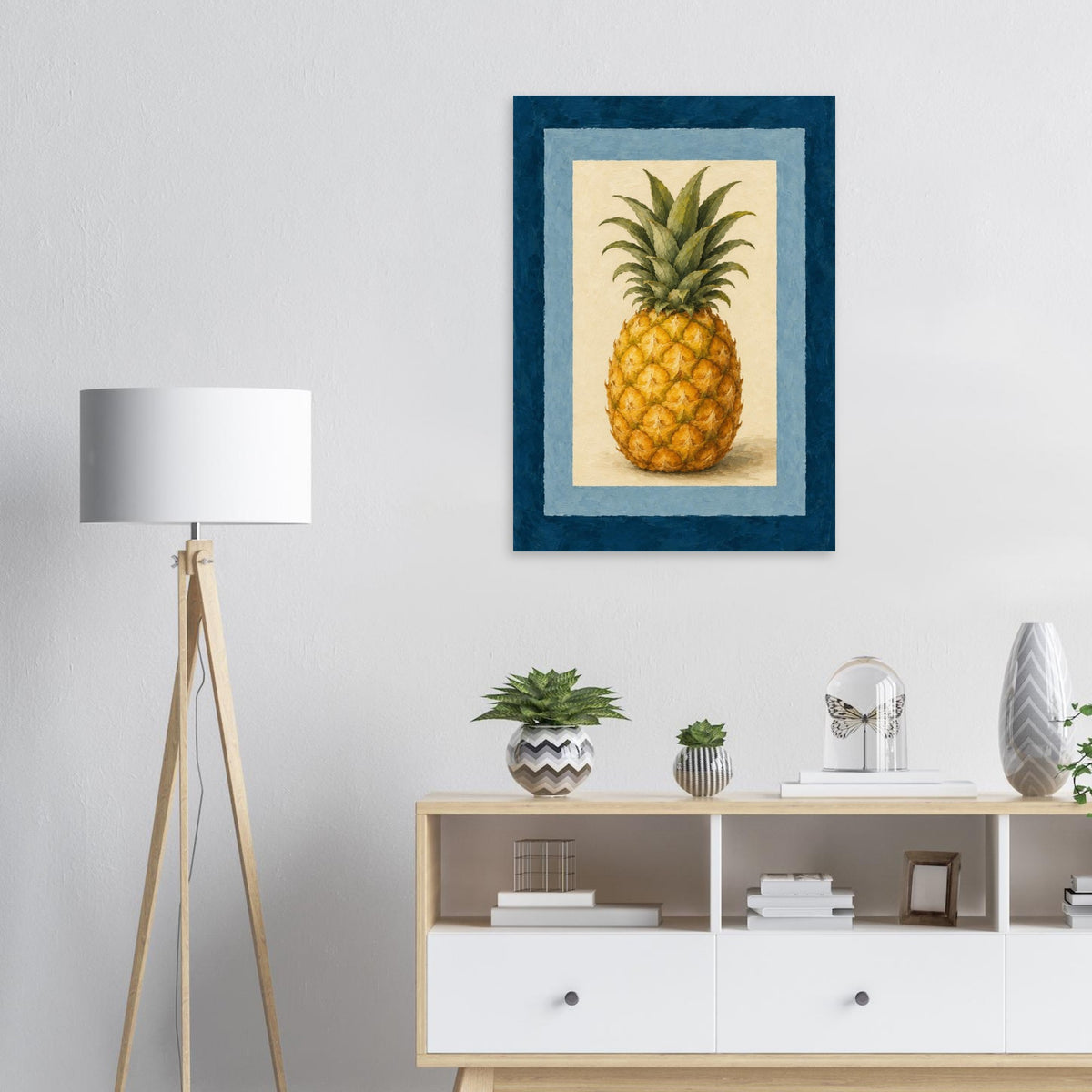 A framed wall art print featuring a pineapple illustration, displayed above a white sideboard in a modern living room setting with a tripod floor lamp and small decor items.