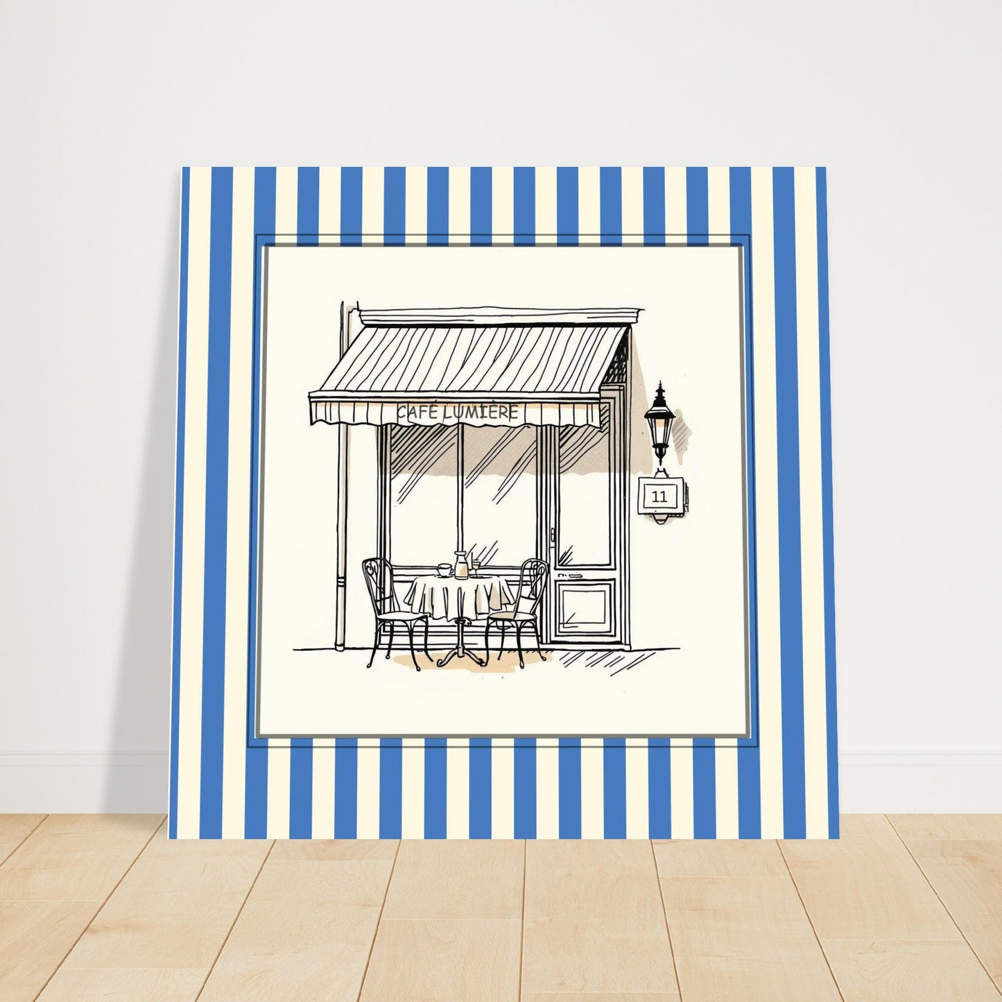 Square French café wall art print with blue striped border leaning against wall on a wooden floor