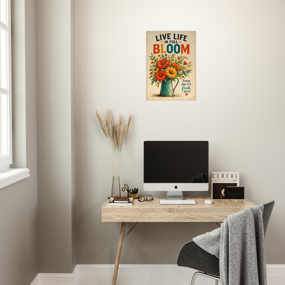 A metal wall art print with the text 'LIVE LIFE IN THE BLOOM' and a colorful floral bouquet hangs above a home office desk setup with a computer, chair, and decorative dried grass in a neutral room.