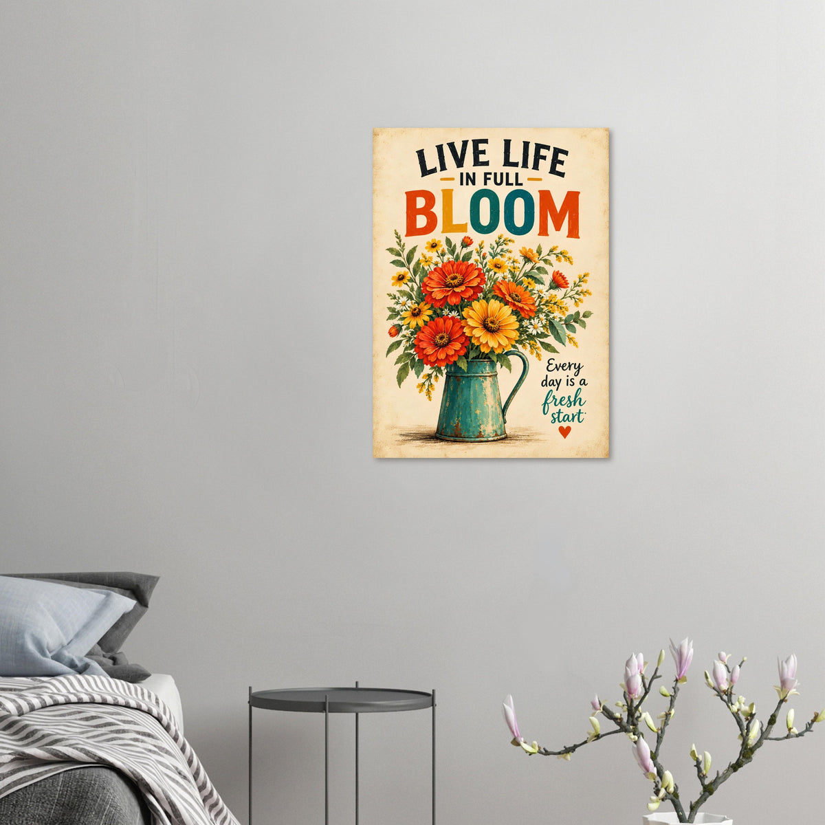 A vertical metal wall art print featuring a colorful floral bouquet in a green pitcher with the text 'LIVE LIFE IN FULL BLOOM' and 'Every day is a fresh start' on a warm beige background, displayed on a light gray wall above a bed and side table.