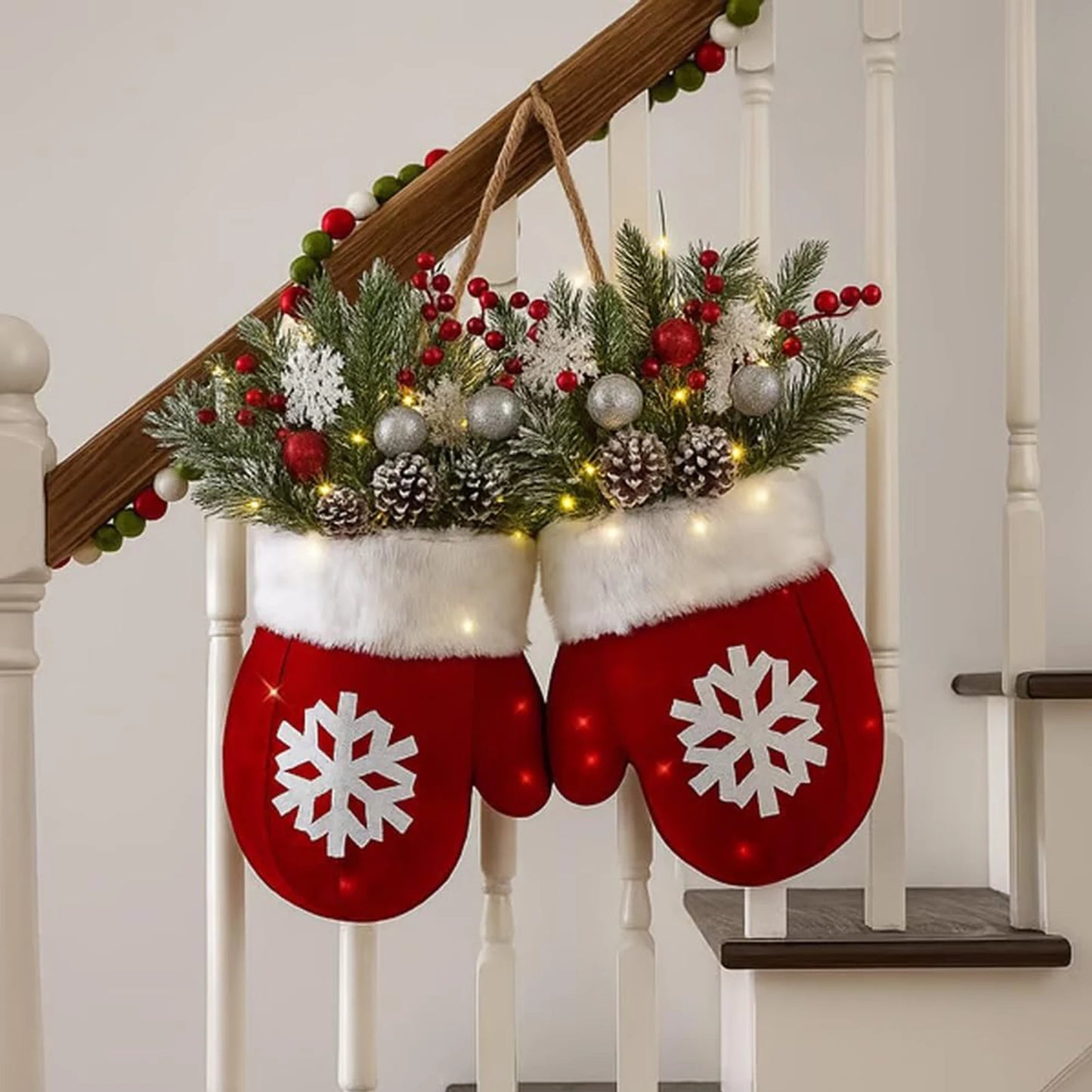 Red mitten-shaped stockings with snowflake designs hanging on a staircase decorated with greenery and lights.