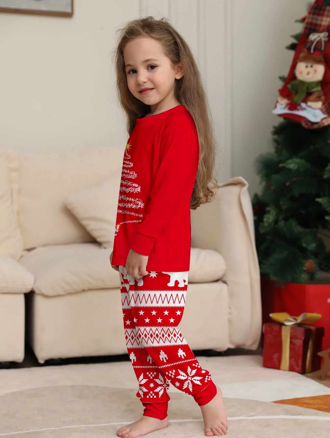 Child wearing red Christmas pajamas in a living room with a tree and presents.