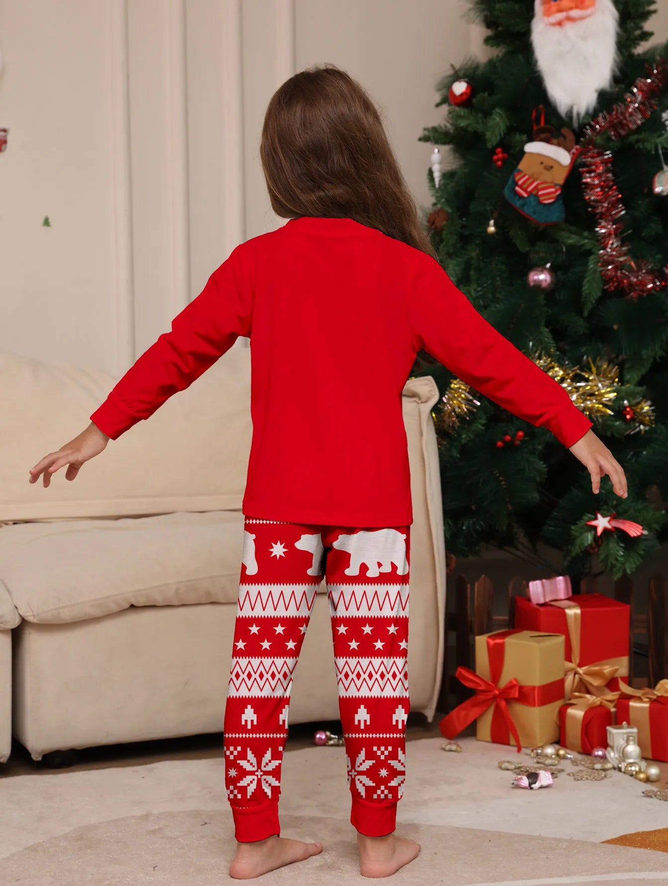 Child wearing red Christmas pajamas with a pattern, standing in front of a decorated Christmas tree and presents.