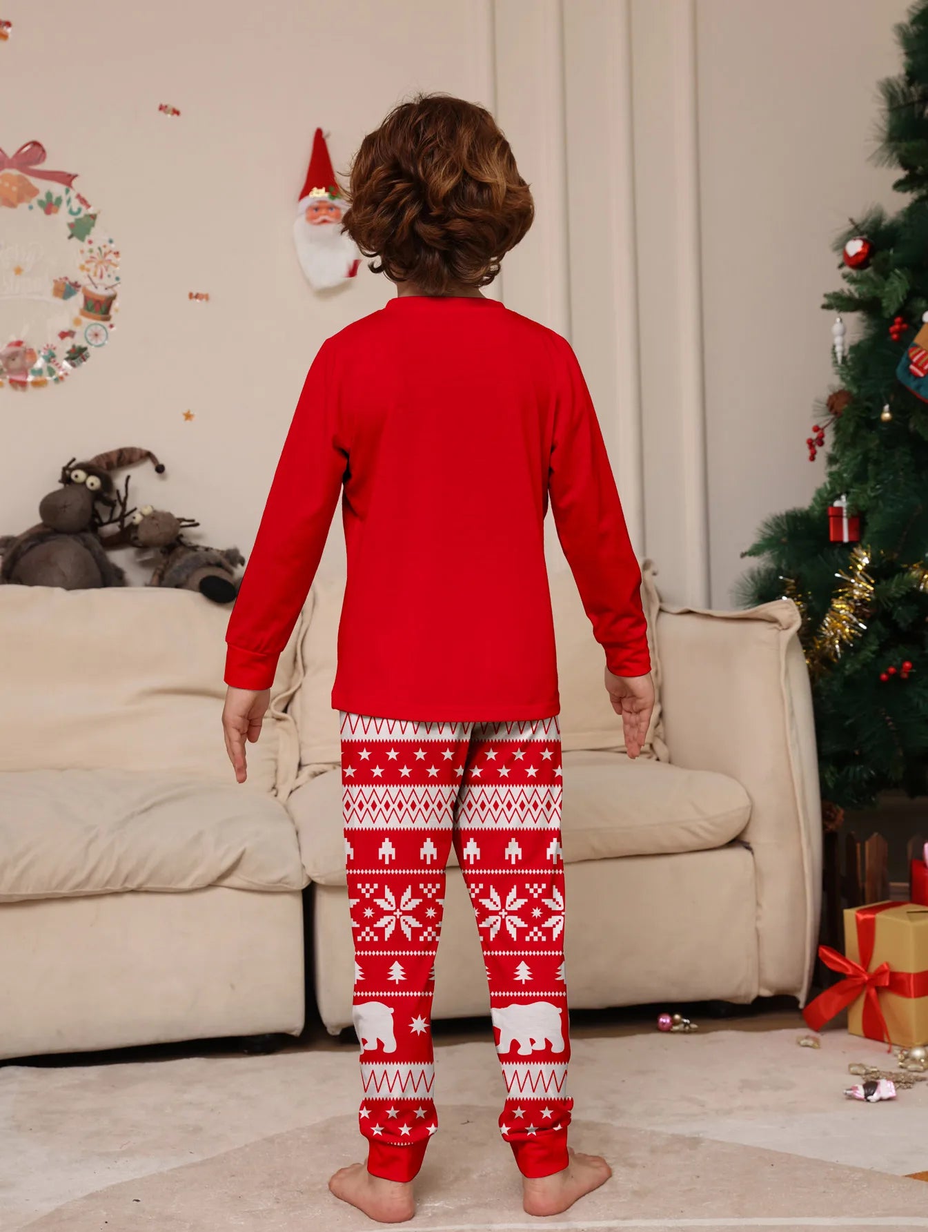 Child wearing red Christmas pajamas with a pattern, standing in a living room with a decorated tree and presents.