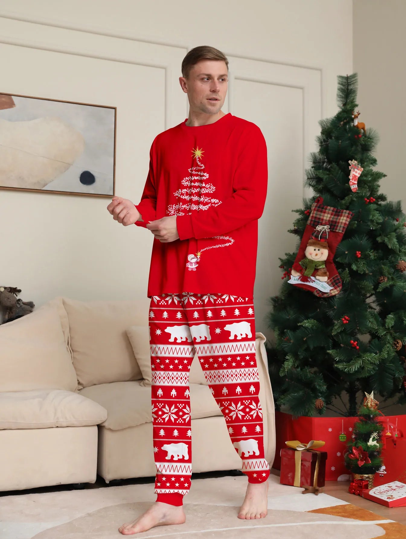 Man wearing a red Christmas-themed pajama set in a living room with a decorated tree and presents.