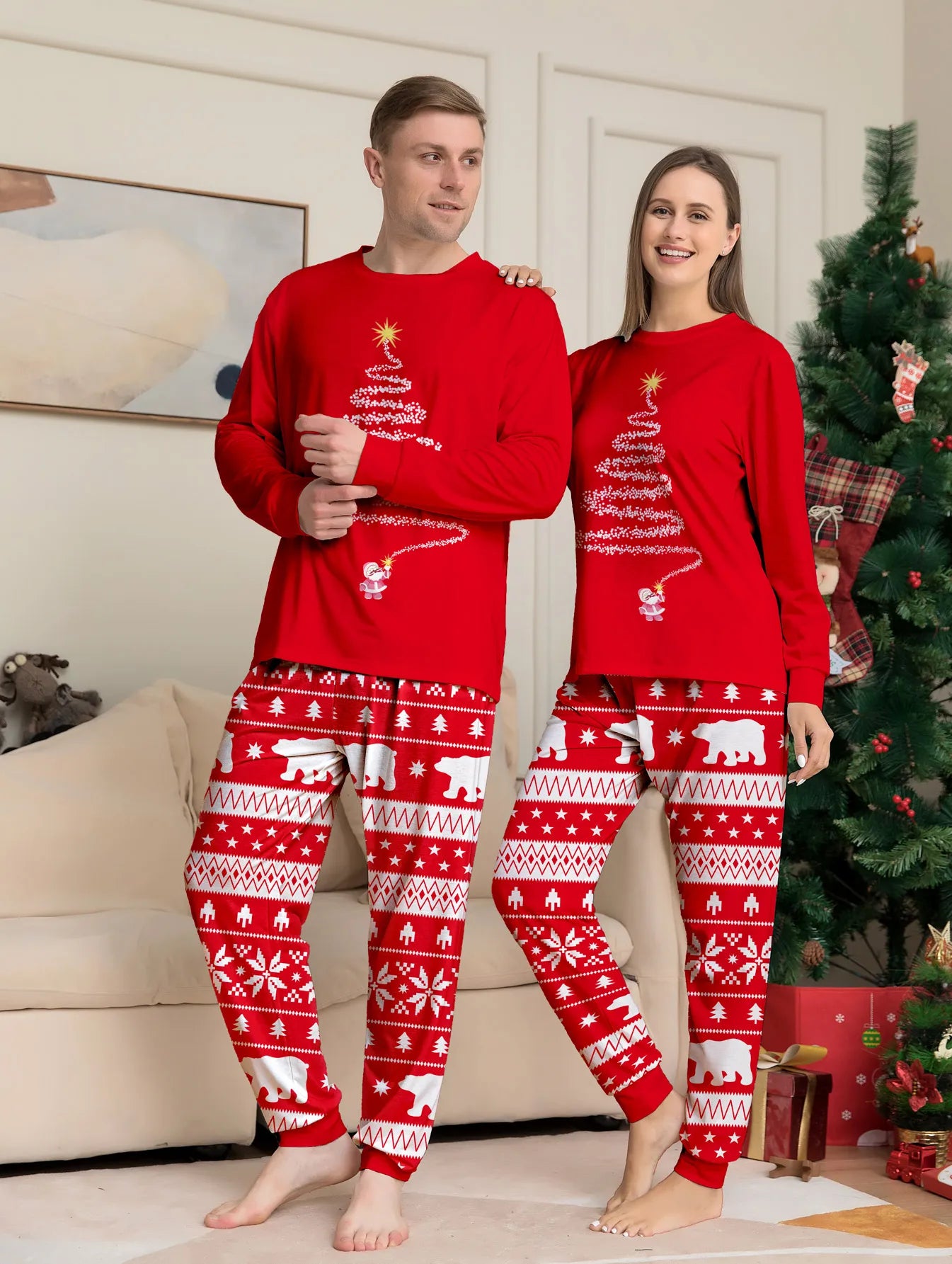 Two people wearing matching red Christmas pajamas in a living room with a decorated tree.