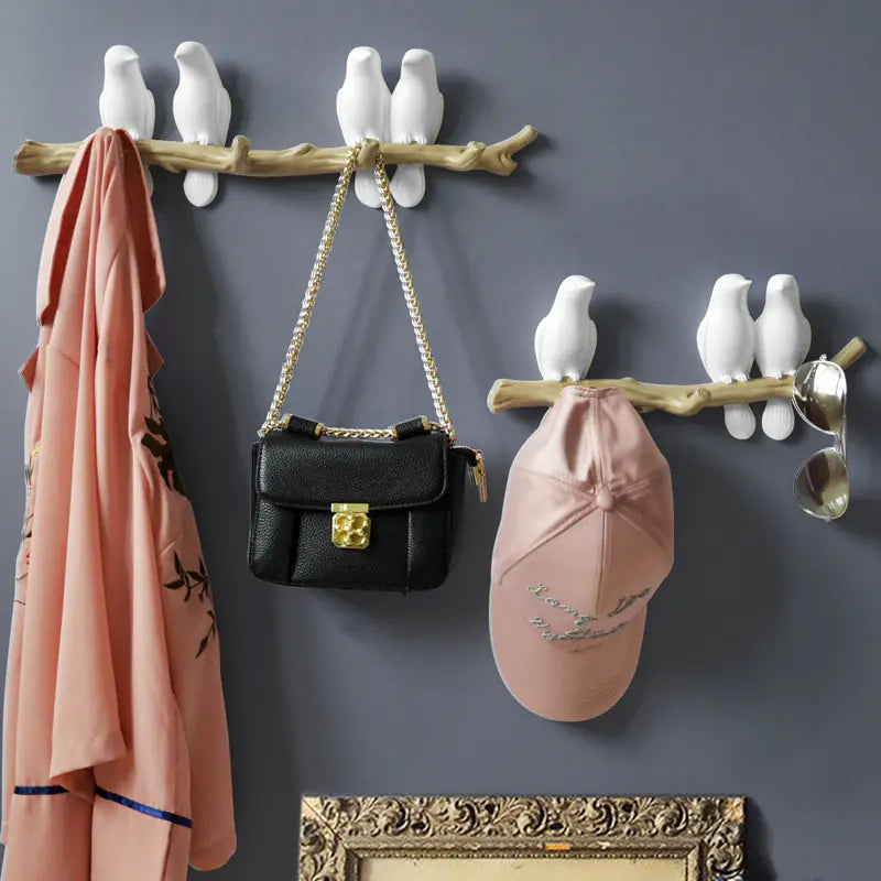 Decorative wall hooks with birds holding a black handbag and pink cap against a gray wall.
