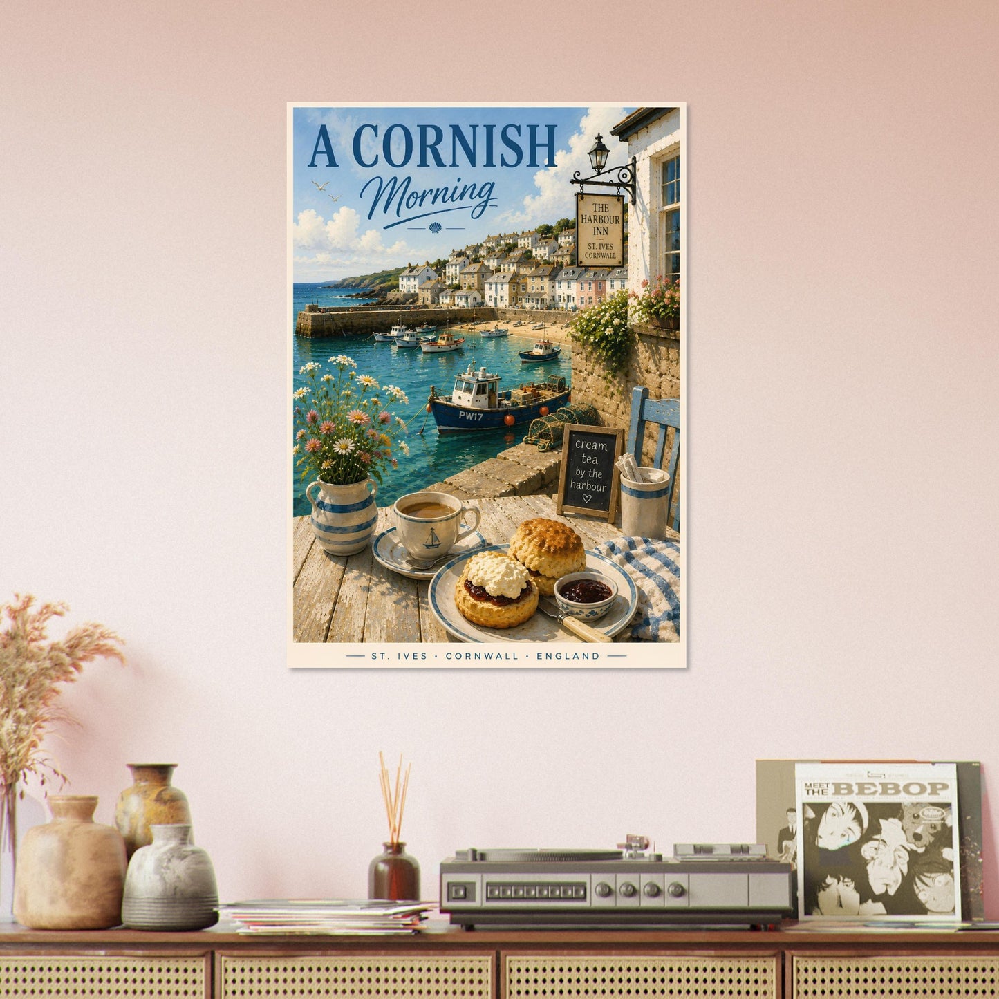 Framed artwork of a Cornish coastal scene with a table setting, framed on a wall above a wooden cabinet.