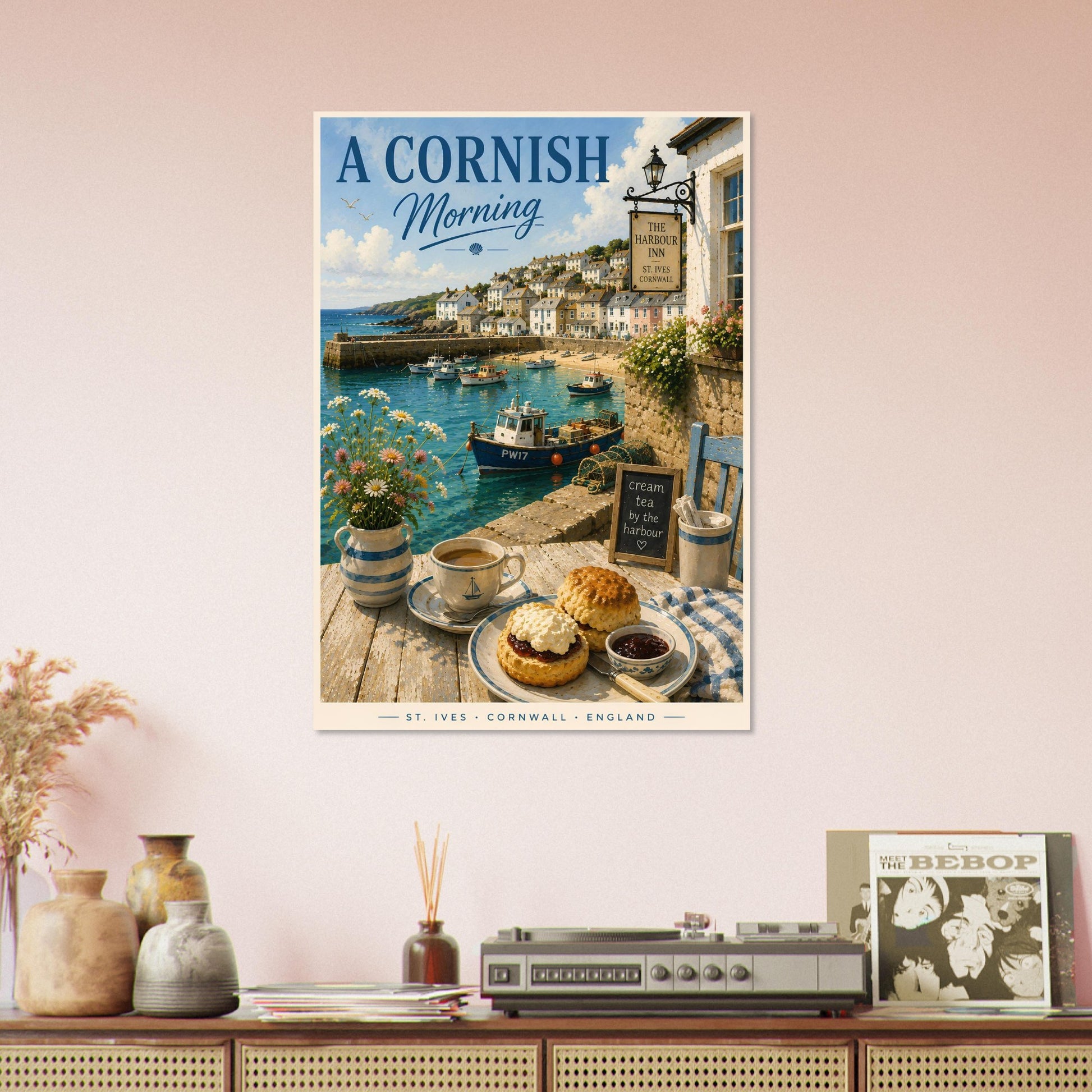 Framed artwork of a Cornish coastal scene with a table setting, framed on a wall above a wooden cabinet.