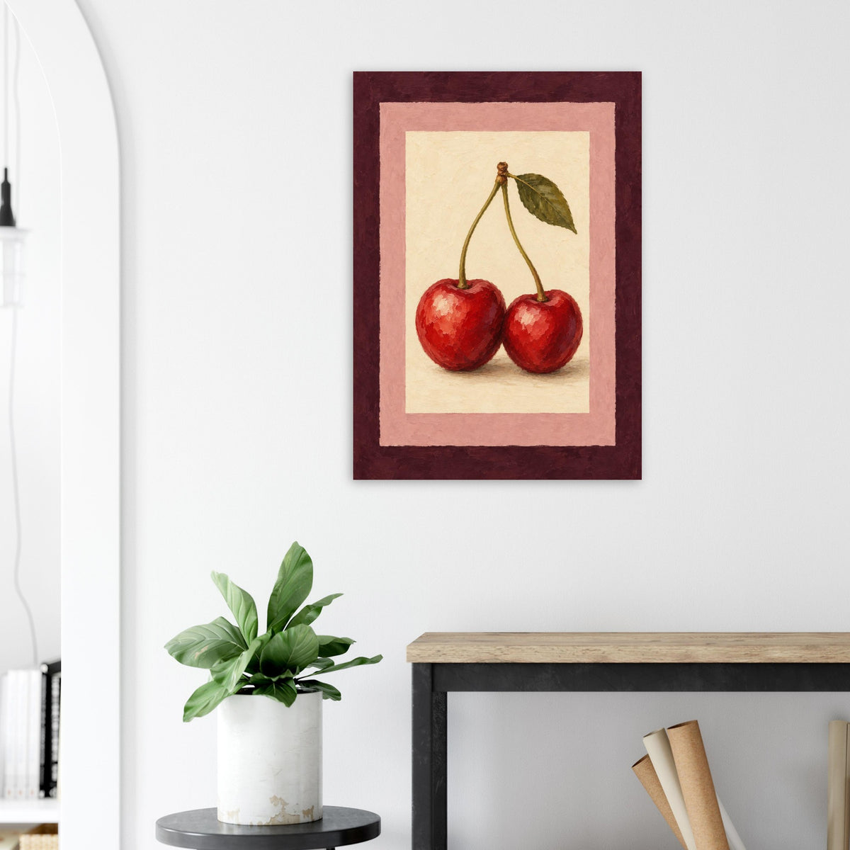 A framed art print featuring a pair of red cherries with green leaves, displayed on a white wall above a console table with a potted plant.