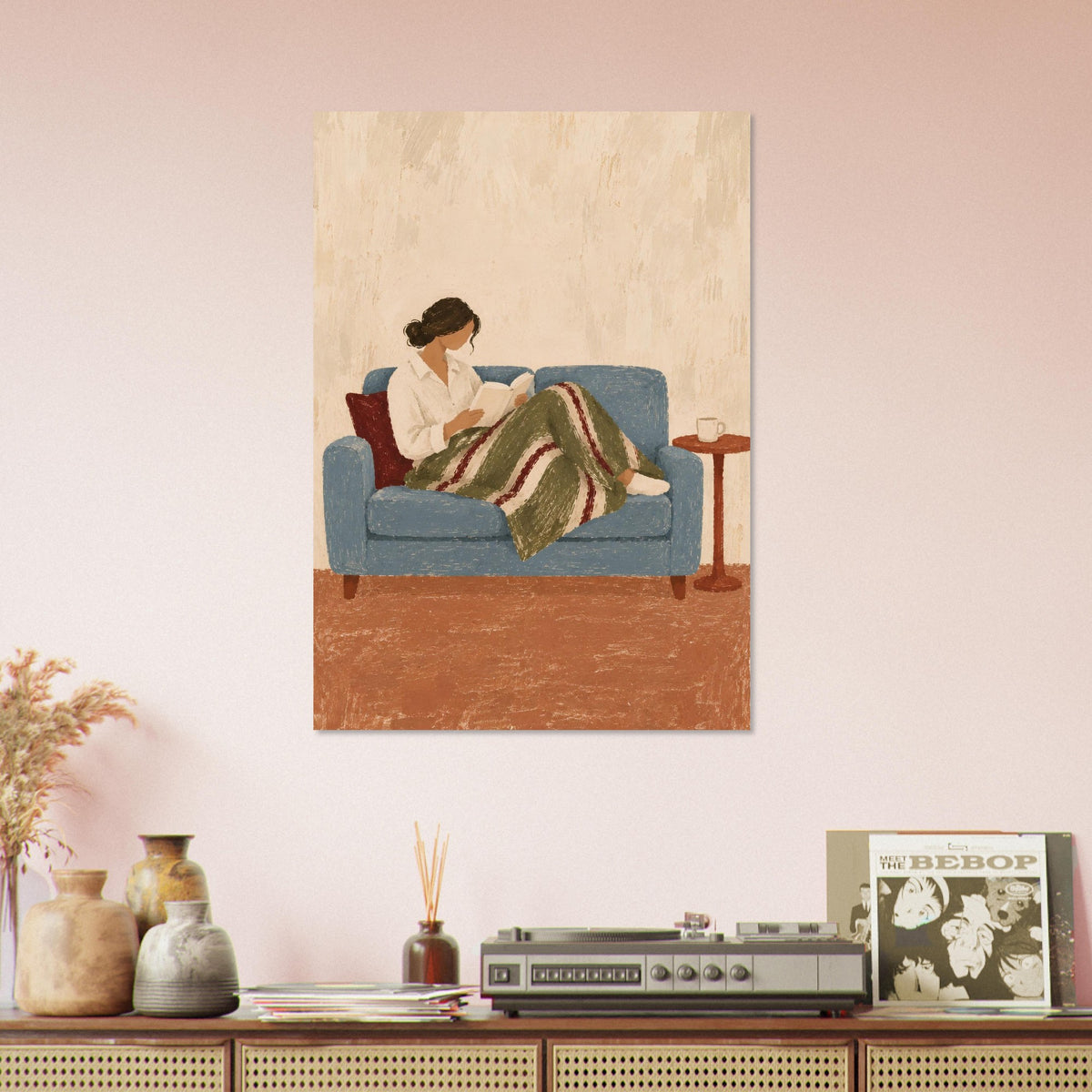 A vertical art print depicting a woman reading on a blue sofa, styled on a light pink wall above a wooden console table with decorative vases, a record player, and a magazine.