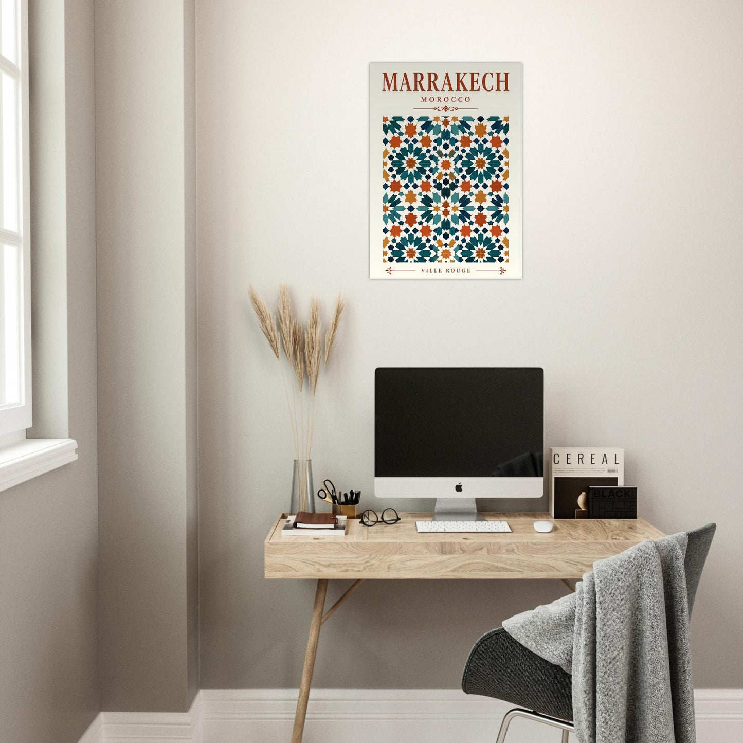 A framed Marrakech Morocco travel poster with a colorful geometric tile pattern hangs on a light beige wall above a wooden desk with a computer monitor, keyboard, and chair in a home office setting.