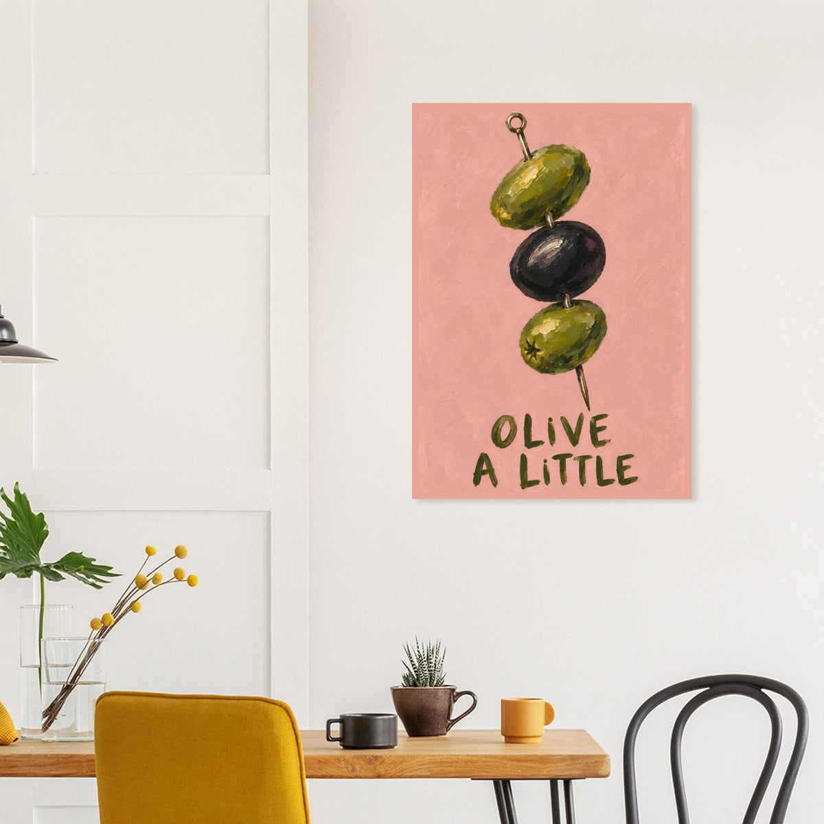 A vertical wall art print featuring three olives on a skewer against a dusty pink background with the text 'OLIVE A LITTLE' in dark green. The print is displayed on a white wall in a modern dining room setting above a wooden table with yellow chairs and small plants.
