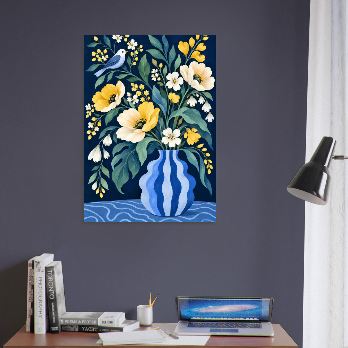 A vertical wall art print featuring a floral bouquet in a blue-and-white striped vase, hung on a dark gray wall above a desk with books, a laptop, and a lamp.