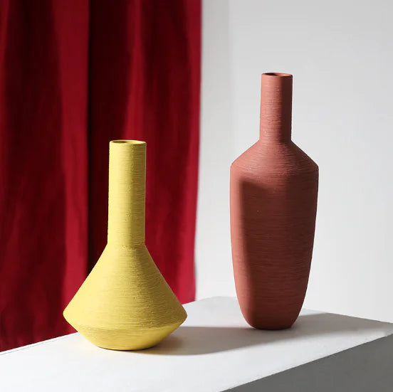 Two ceramic vases, one yellow and one terracotta, on a white surface with a red curtain in the background.