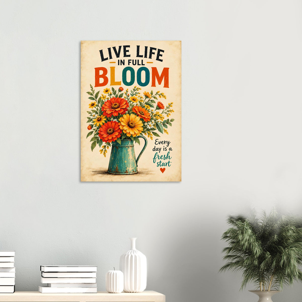 A vertical metal wall art print featuring a floral bouquet in a teal pitcher with the text 'LIVE LIFE IN FULL BLOOM' and 'Every day is a fresh start' on a light gray wall above a console table with books and a plant.