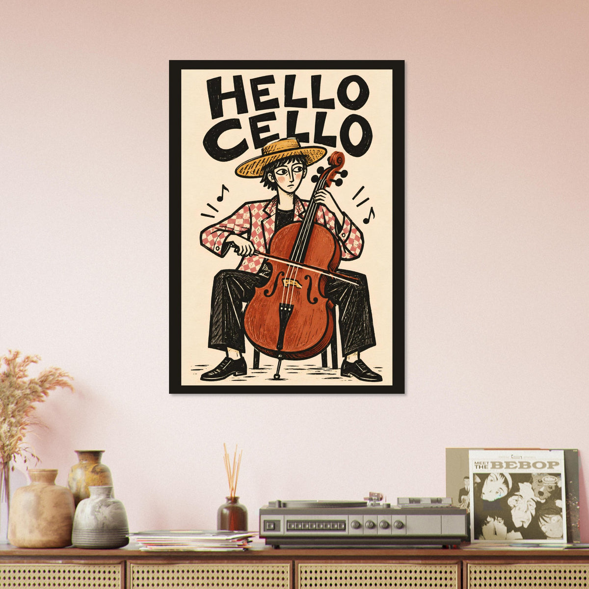 A framed art print featuring a retro-style illustration of a cellist with the text 'HELLO CELLO' on a cream background, hanging on a light pink wall above a wooden console table with decorative vases, a record player, and a magazine.