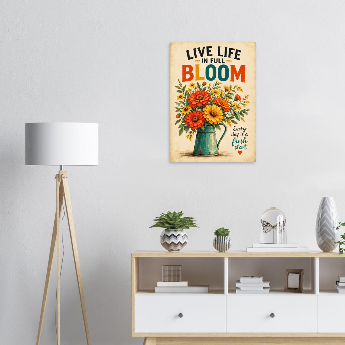A vertical metal wall art print featuring a vintage-style illustration of sunflowers and a teal pitcher with the text 'LIVE LIFE IN FULL BLOOM' and 'Every day is a fresh start', displayed on a light gray wall above a white and light wood console cabinet in a styled room setting.