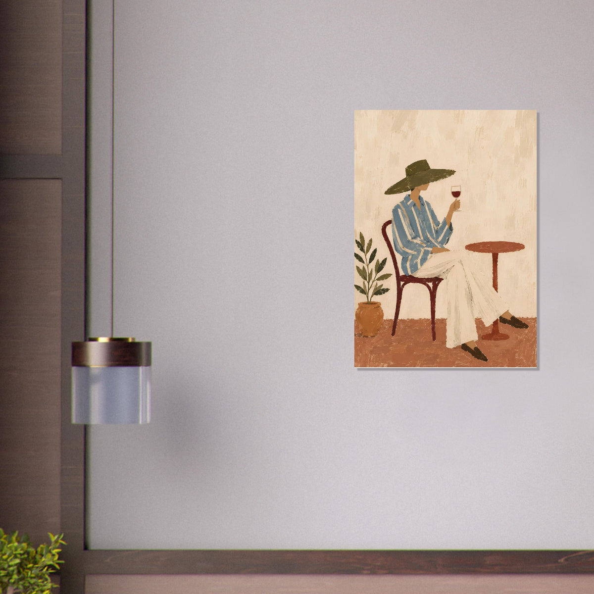 A vertical art print depicting a woman in a wide-brimmed hat sitting at a table with a glass of red wine, displayed on a light gray wall next to a modern wall lamp.