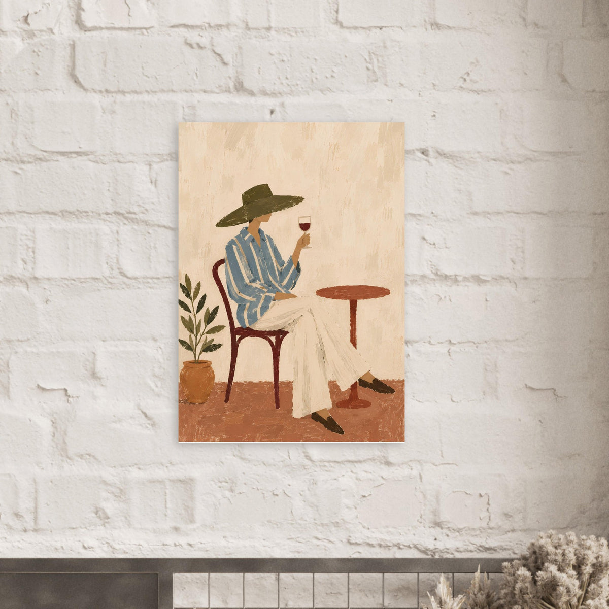 A vertical art print featuring a stylized illustration of a person in a wide-brimmed hat, striped shirt, and cream trousers sitting at a small table and holding a glass of red wine. The print is mounted on a white brick wall.
