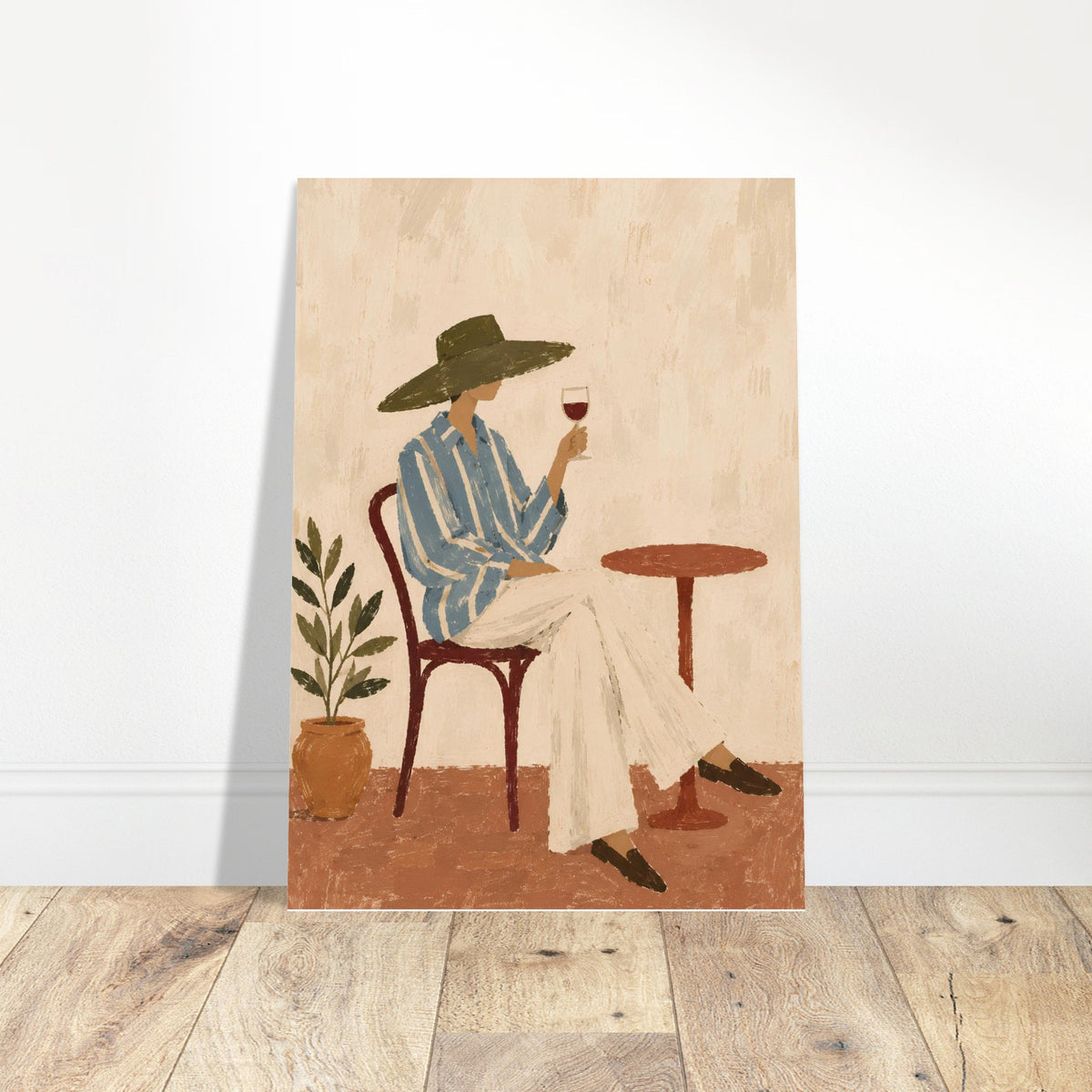 A vertical art print featuring a stylized illustration of a woman in a blue striped shirt and wide-brimmed hat sitting at a small table with a glass of red wine, leaning against a white wall on a light wood floor.