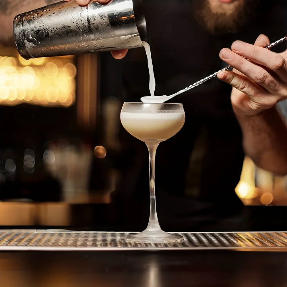 Barista pouring a creamy liquid into a glass cup with a blurred bar setting in the background