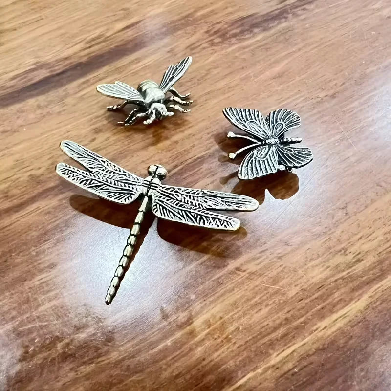 Three metallic dragonfly and insect-shaped ornaments arranged on a wooden tabletop.