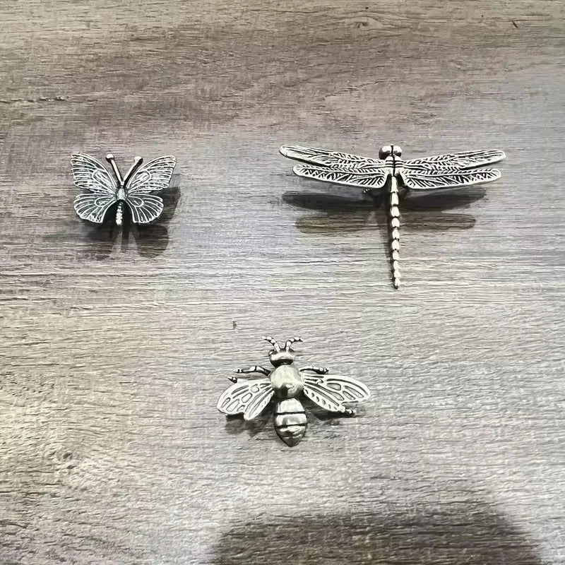 Three small metallic insect cecorativew pieces, including a butterfly, dragonfly, and bee, displayed on a gray wood-grain surface.