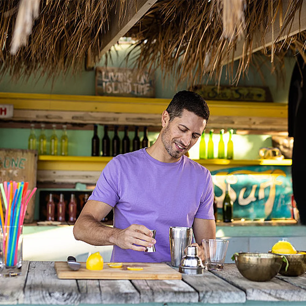 Man in a purple shirt preparing a drink at a bar counter with various bottles and ingredients.