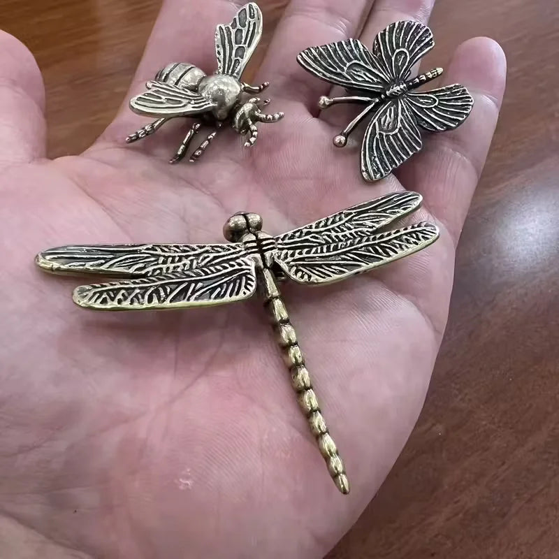 Three metallic insect-shaped decorative pieces, including a dragonfly and two bees, displayed on an open hand over a wooden surface.