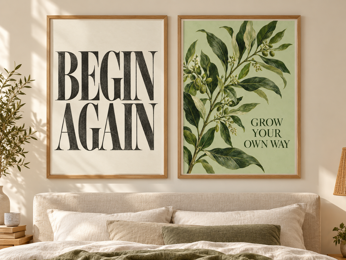 Two framed wall art prints displayed above a bed in a softly lit bedroom. The left print has large black text reading “BEGIN AGAIN” on a cream background, and the right print shows green botanical leaves with the text “GROW YOUR OWN WAY” on a pale green background.