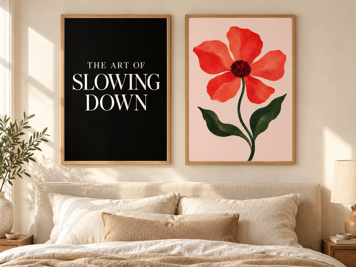 Two framed wall art prints displayed above a bed in a softly lit bedroom. The left print has a black background with white text reading “THE ART OF SLOWING DOWN,” and the right print shows a large red-orange flower with green leaves on a pale pink background.